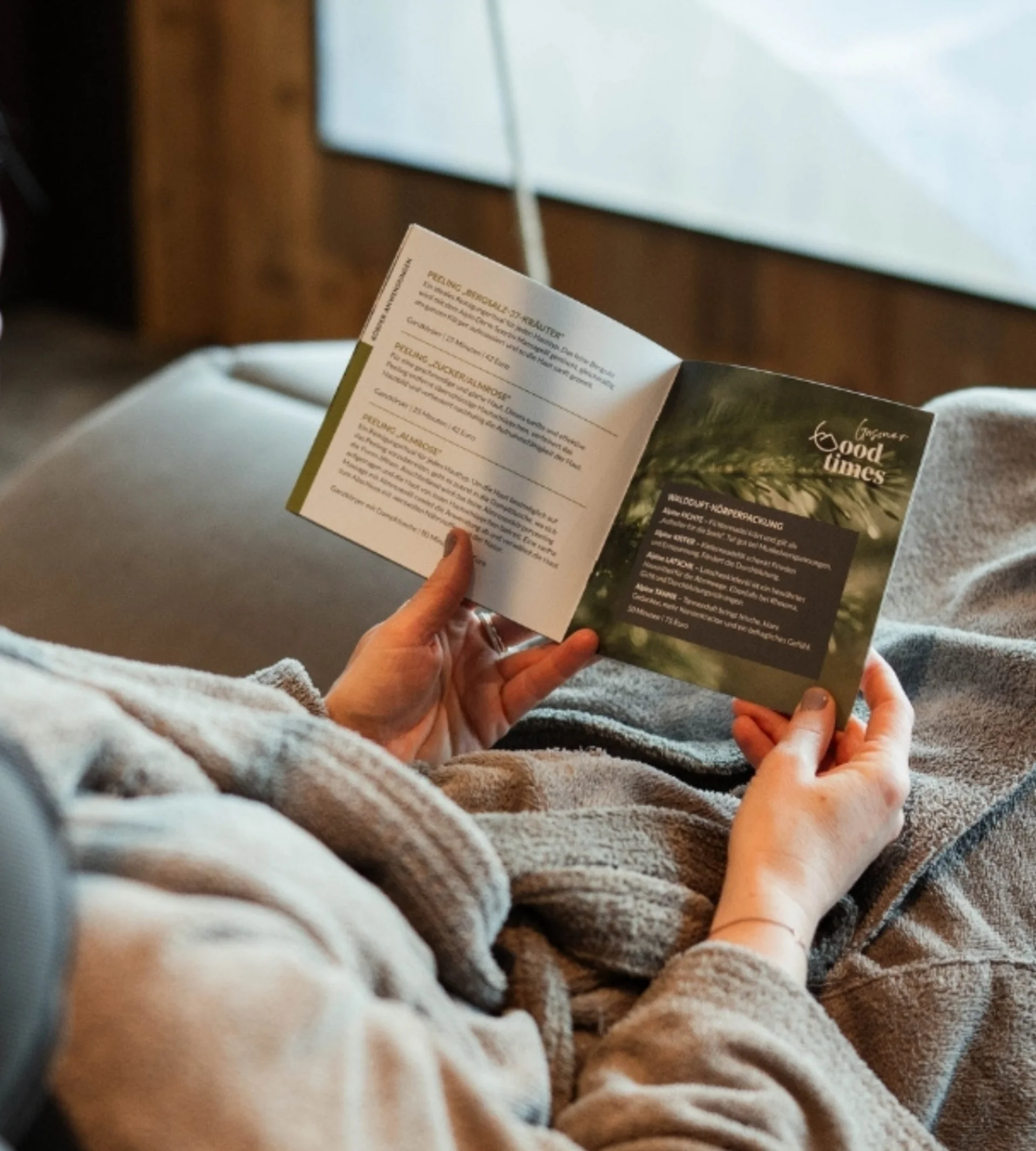 Wellness Person liest entspannt Broschüre auf Sofa mit Decke und Blick zum Fenster