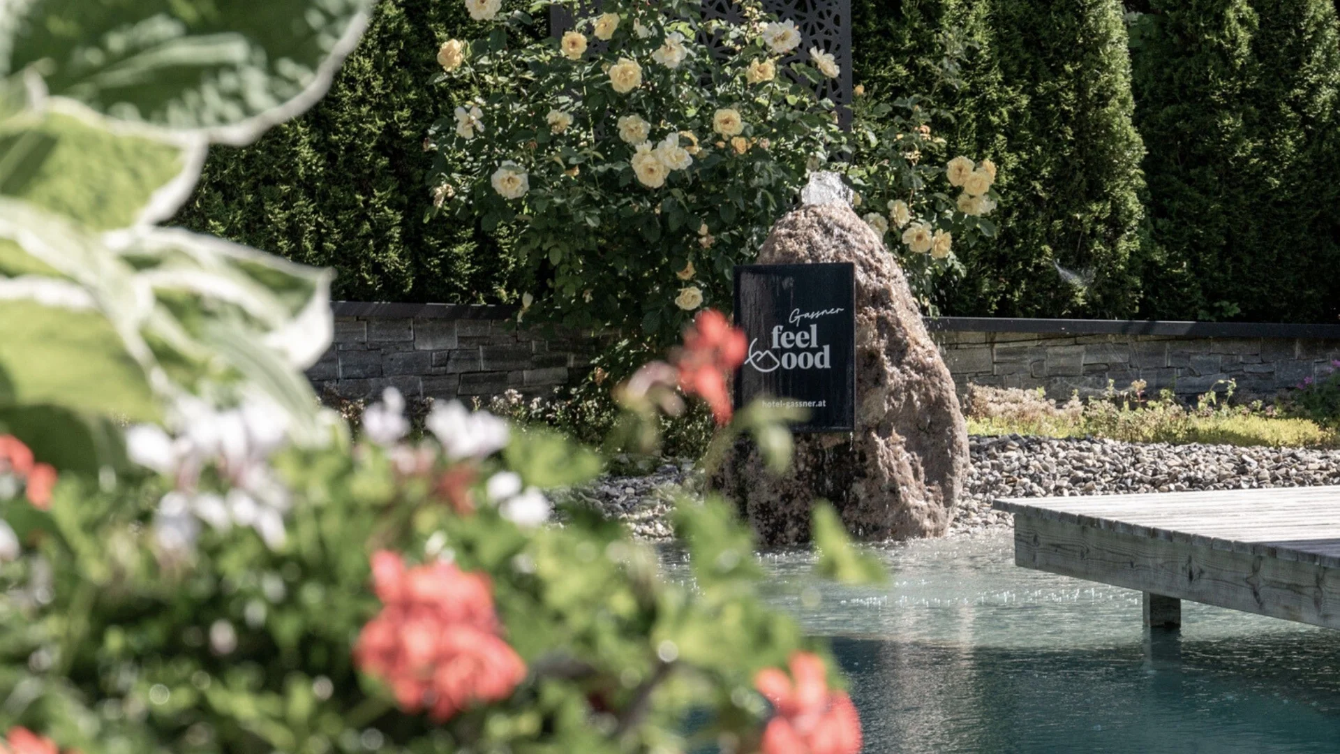Saunawelt Gartenteich mit Blumen, Steinskulptur und Holzsteg im sonnigen Garten
