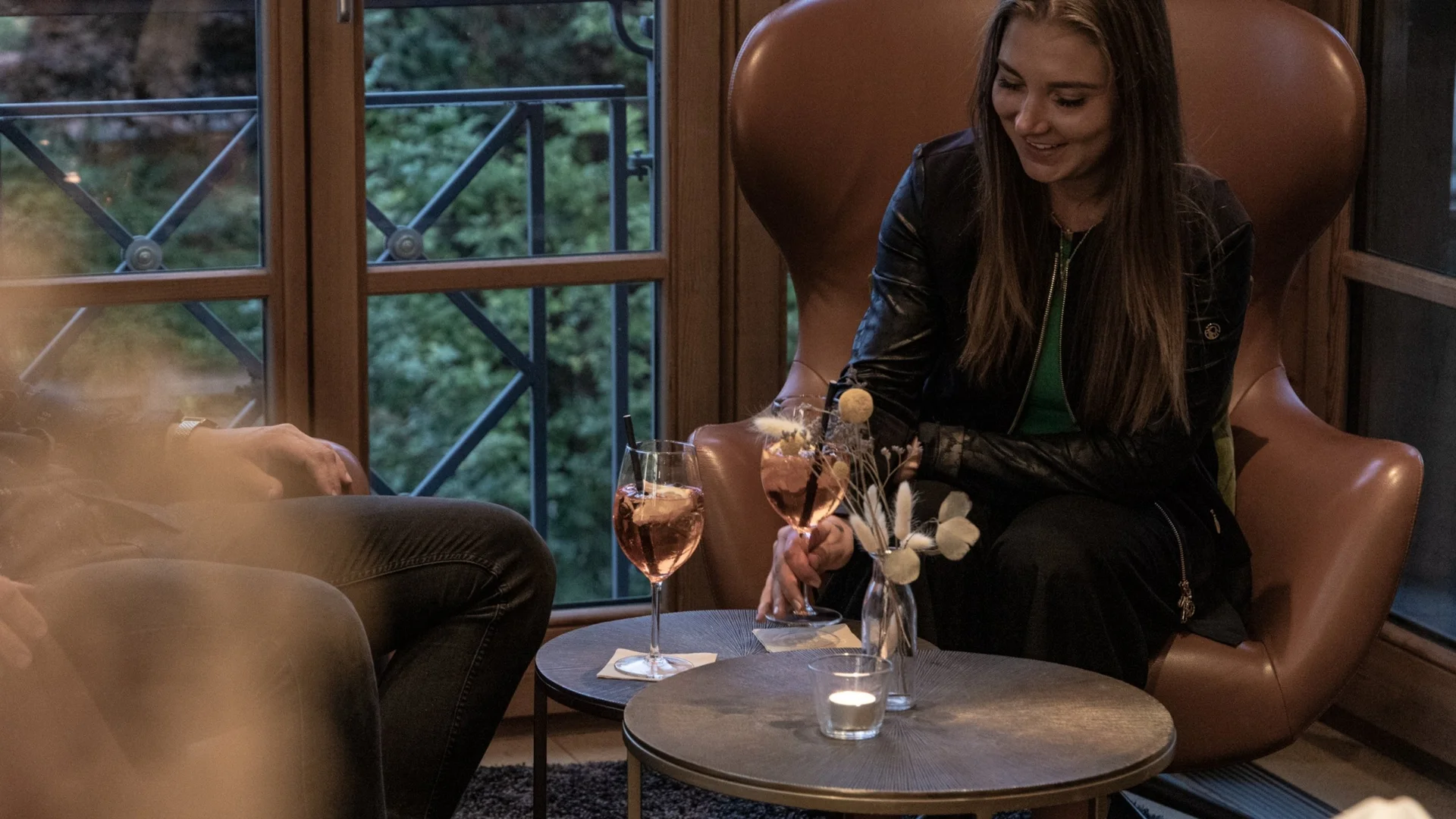 WORK FOR US Frau in Lederjacke sitzt mit Getränken an Tisch in gemütlicher Lounge