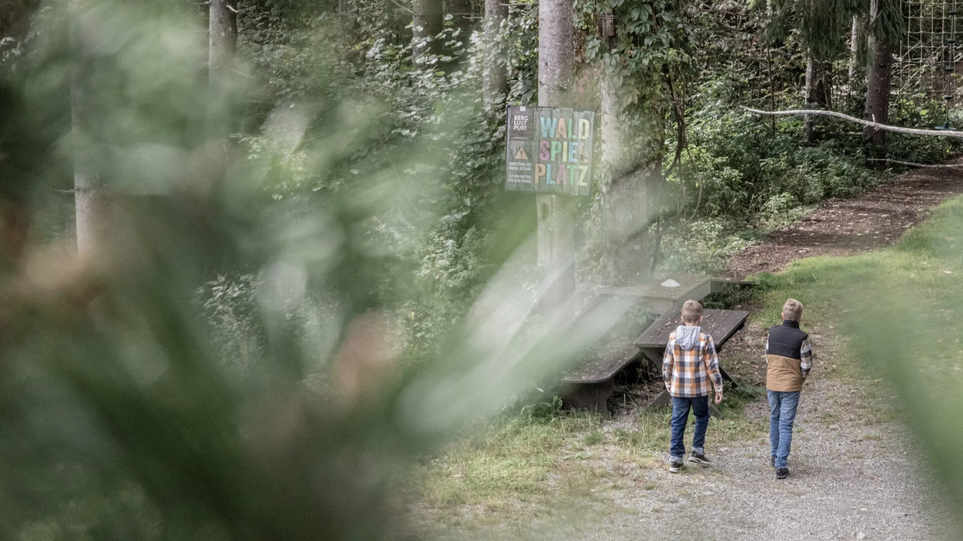 Baumhaus Zwei Jungen gehen auf einem Waldweg an einem Schild für Waldspielplatz vorbei