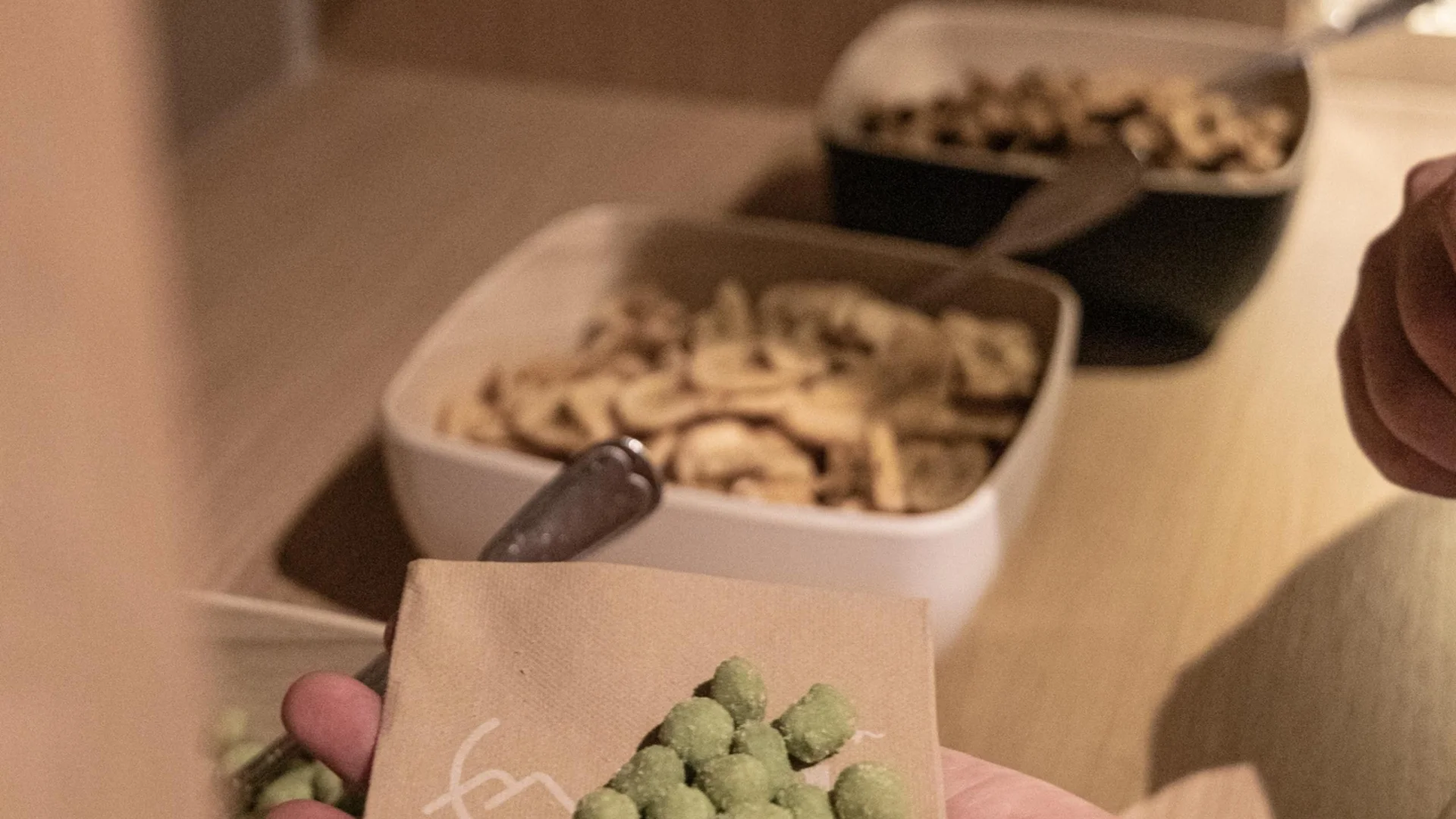 Saunawelt Hand hält grüne Wasabi-Erbsen auf Serviette vor Schalen mit Snacks