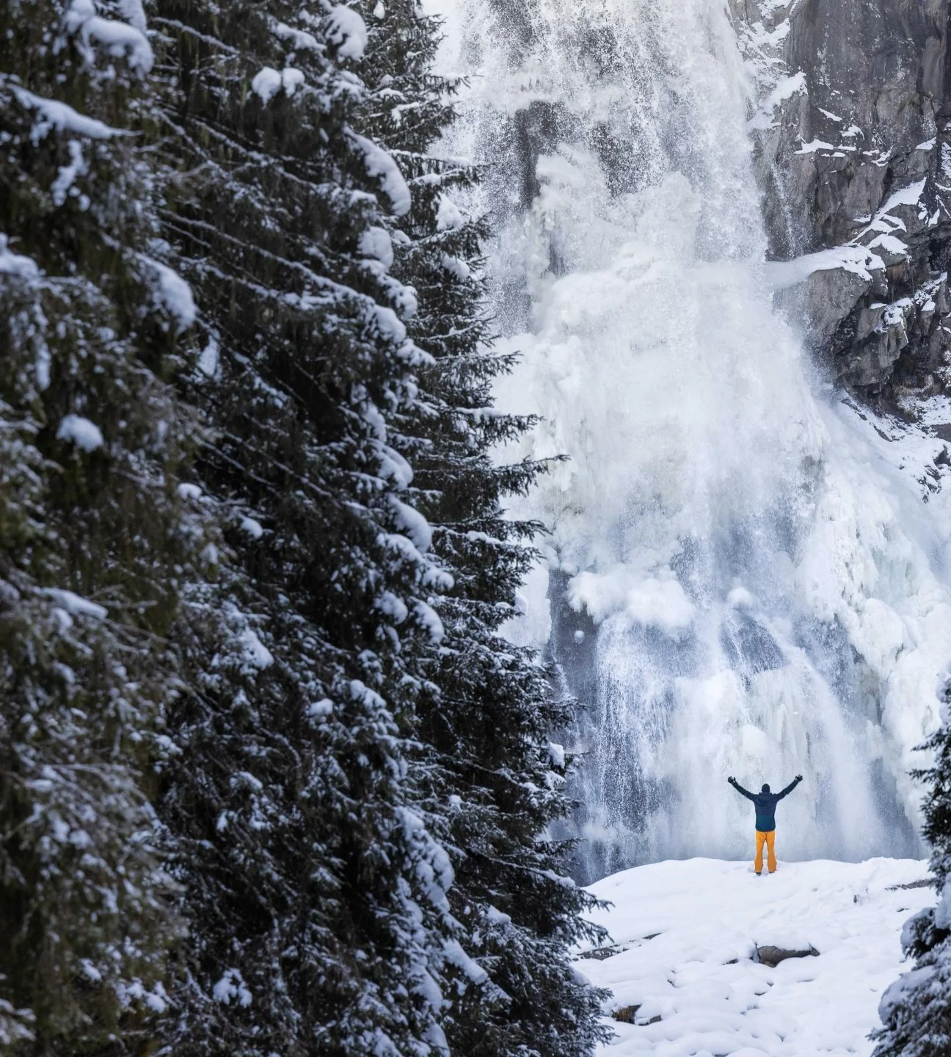 Winterwandern Person mit erhobenen Armen vor einem verschneiten gefrorenen Wasserfall umgeben von Kiefern