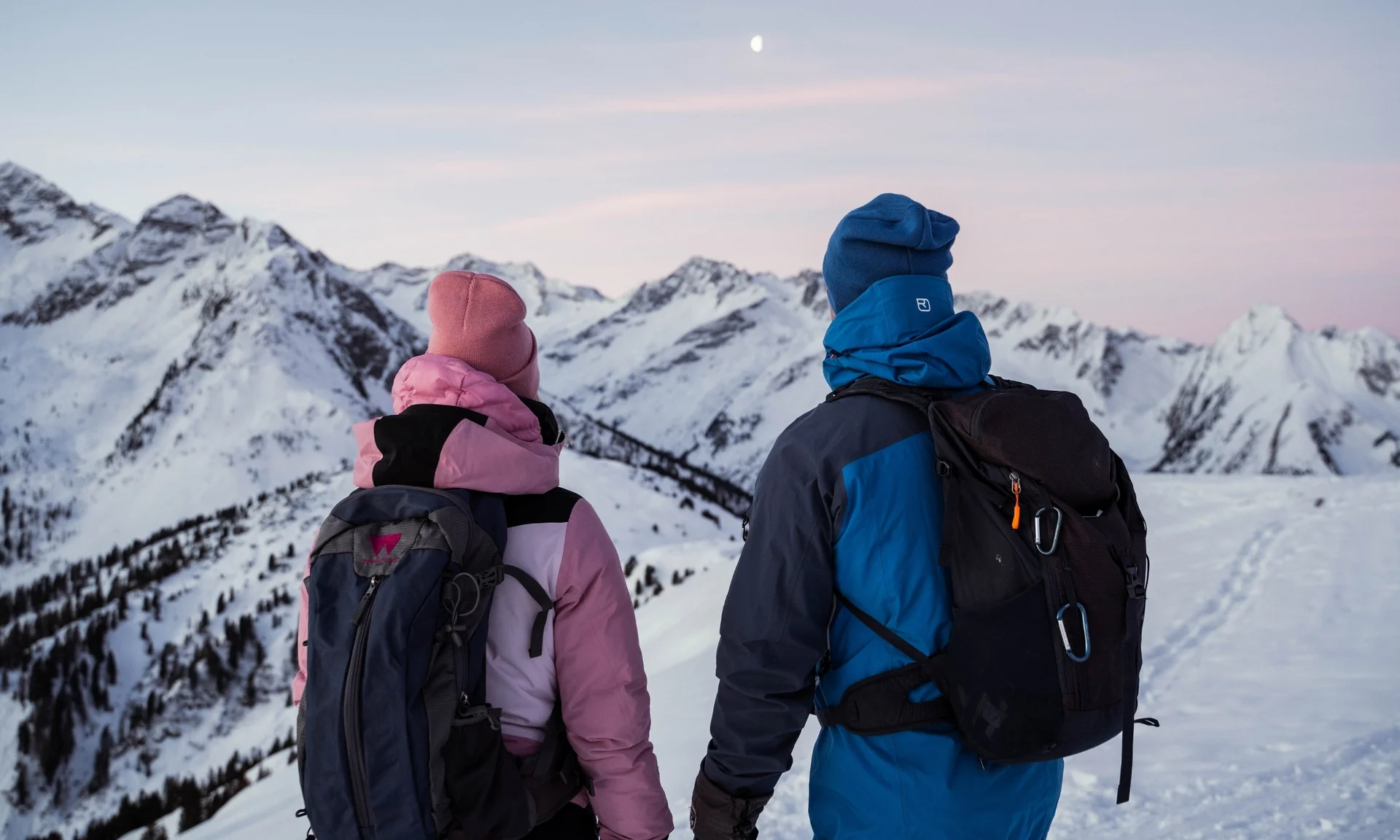 Home Two hikers in winter gear looking at snow-covered mountains at dusk