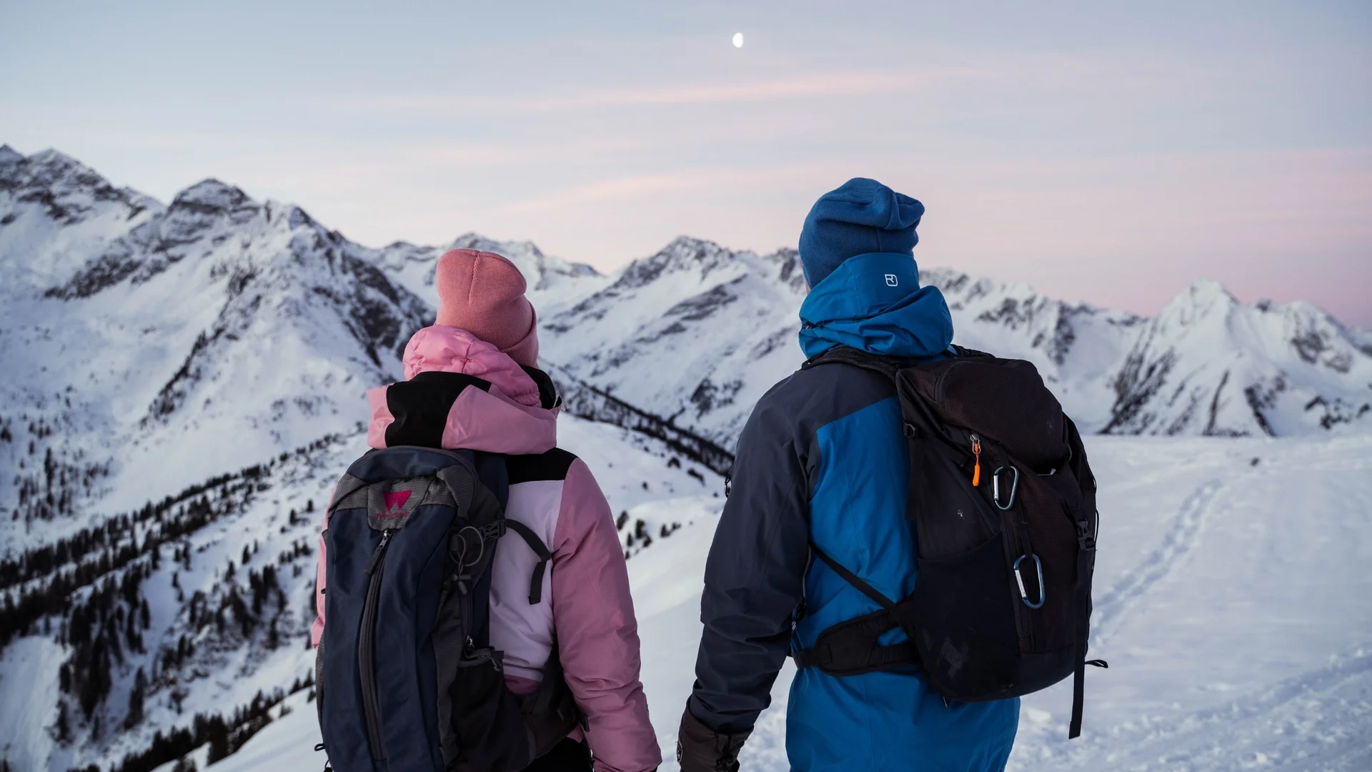 Wanderhotels Empathieprogramm Zwei Wanderer in Winterkleidung blicken auf verschneite Berge bei Dämmerung