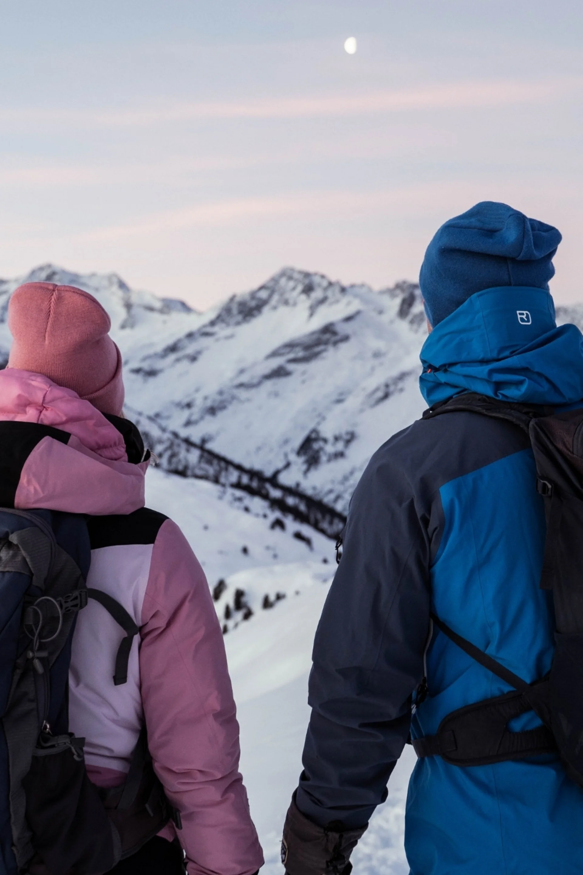 Inklusivleistungen Zwei Wanderer in Winterkleidung blicken auf verschneite Berge bei Dämmerung