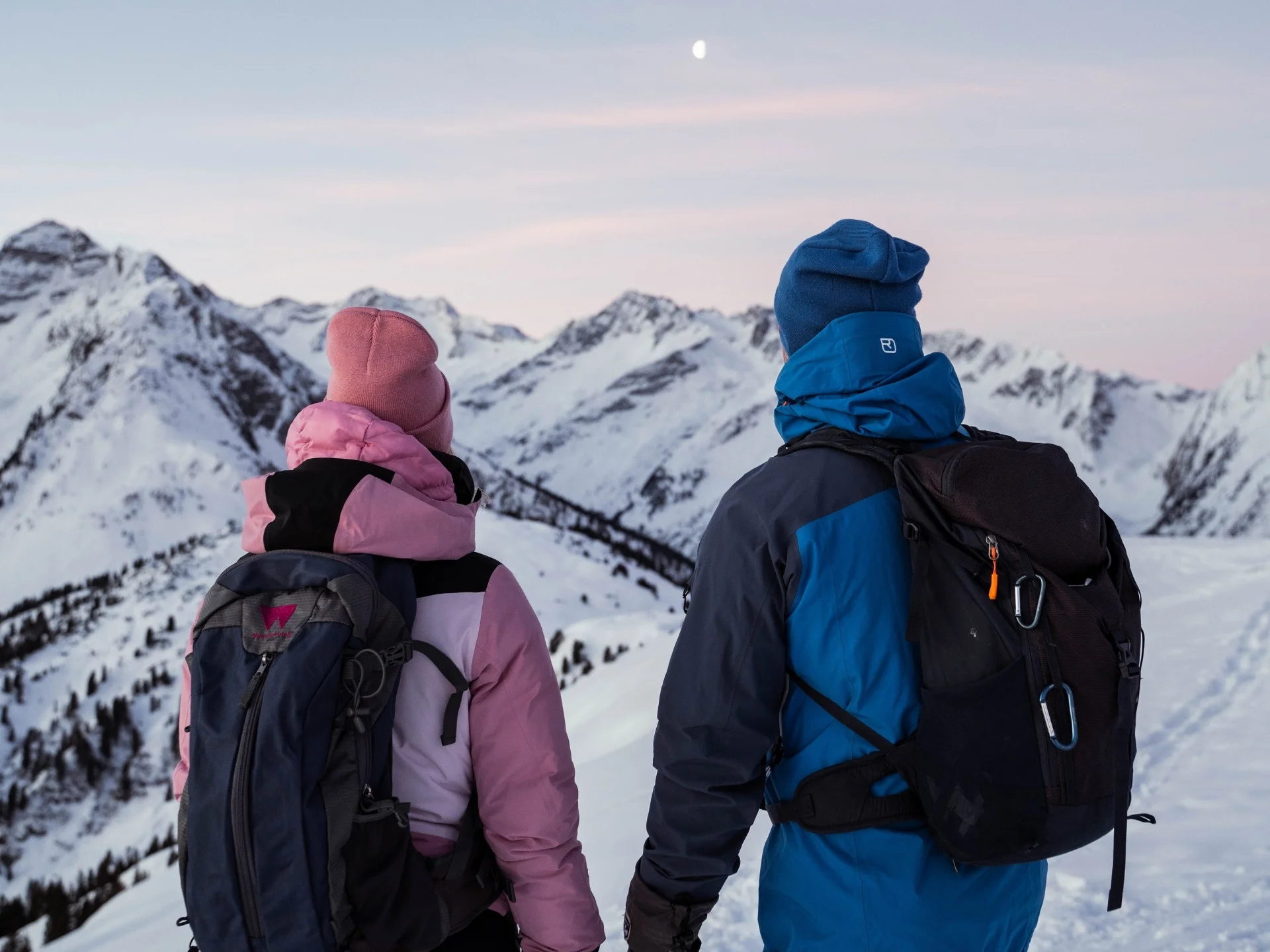 Wanderservice Zwei Wanderer in Winterkleidung blicken auf verschneite Berge bei Dämmerung