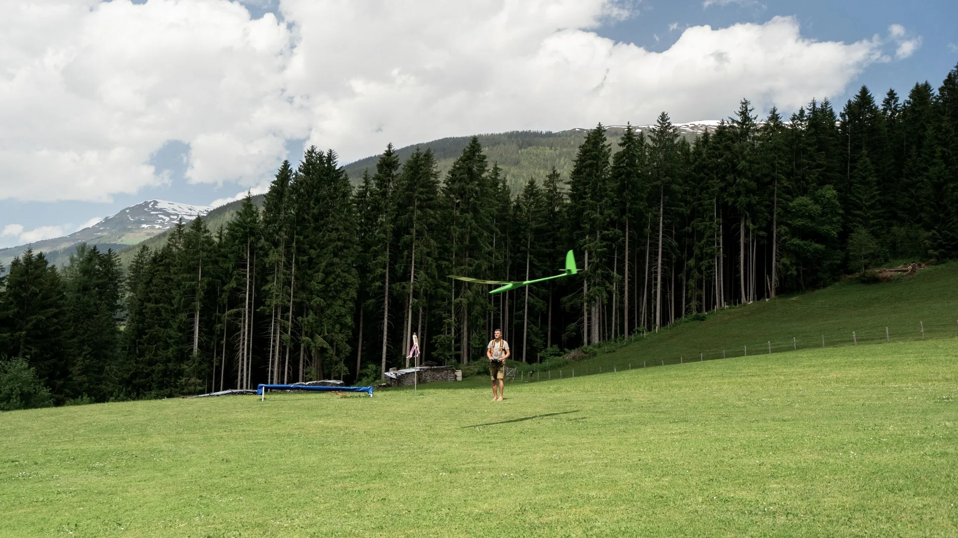 Modell- und Hangfliegen Mann fliegt grünes Modellflugzeug auf Wiese vor Wald und Bergen