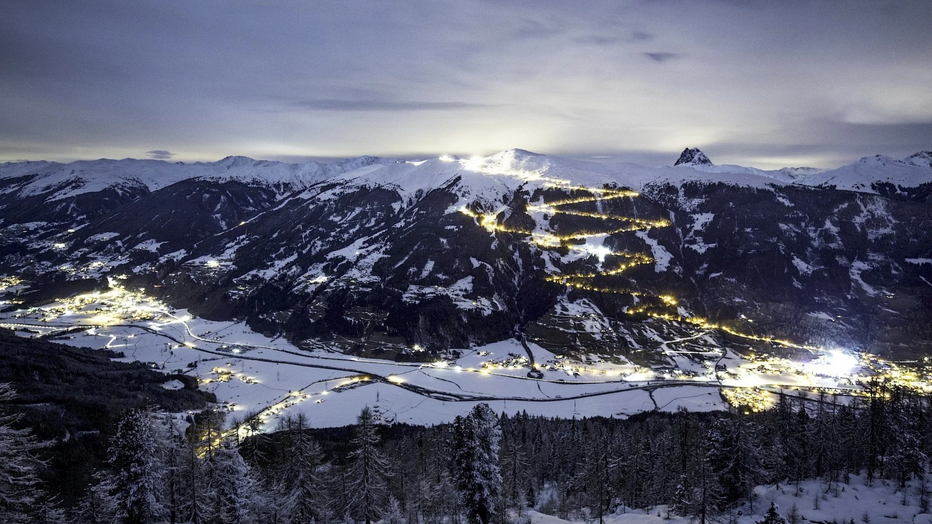 Rodeln Schneebedeckte Berge mit beleuchteten Skiwegen und einem verschneiten Tal bei Nacht