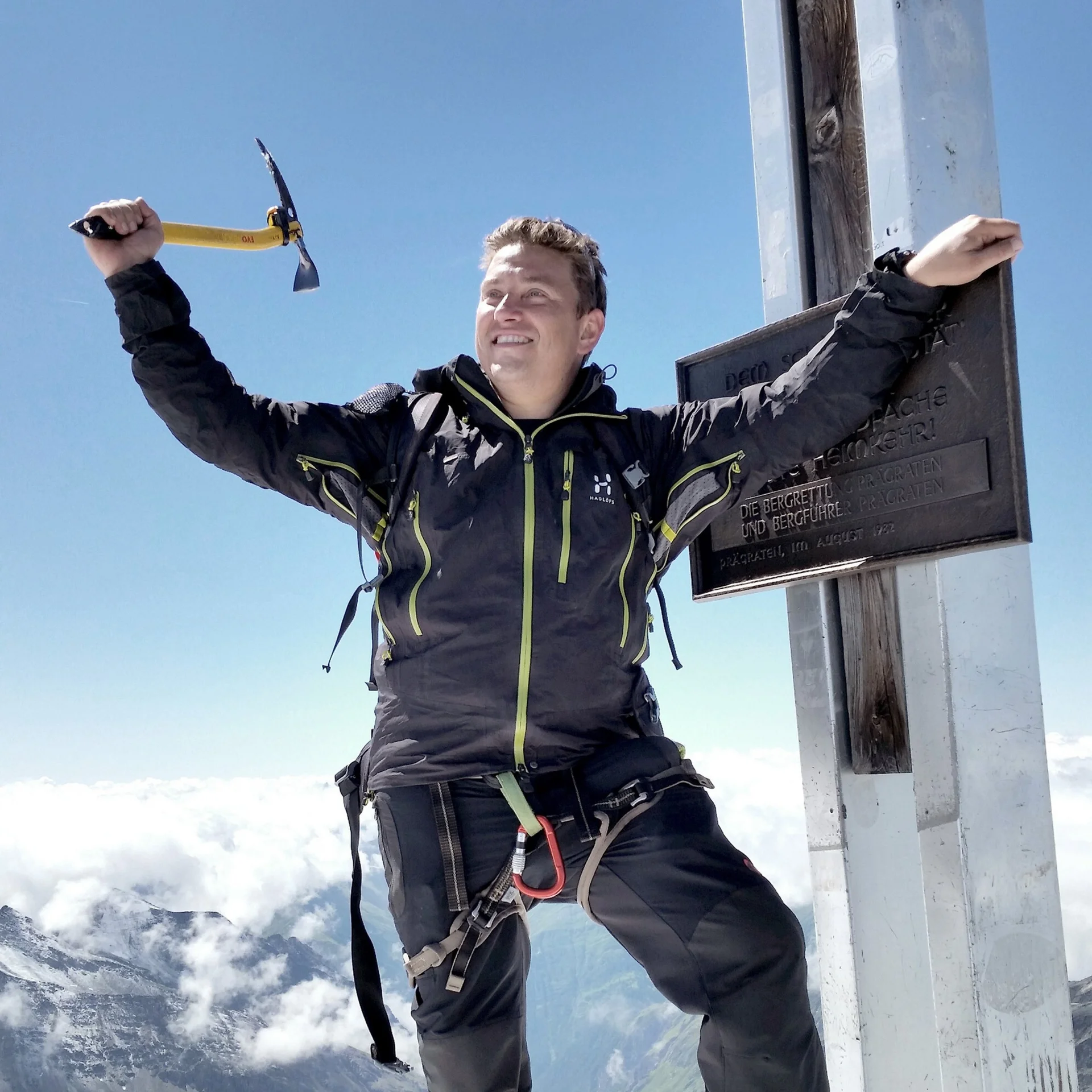 Großvenediger Glücklicher Bergsteiger mit Eispickel am Gipfelkreuz auf bergigem Hintergrund