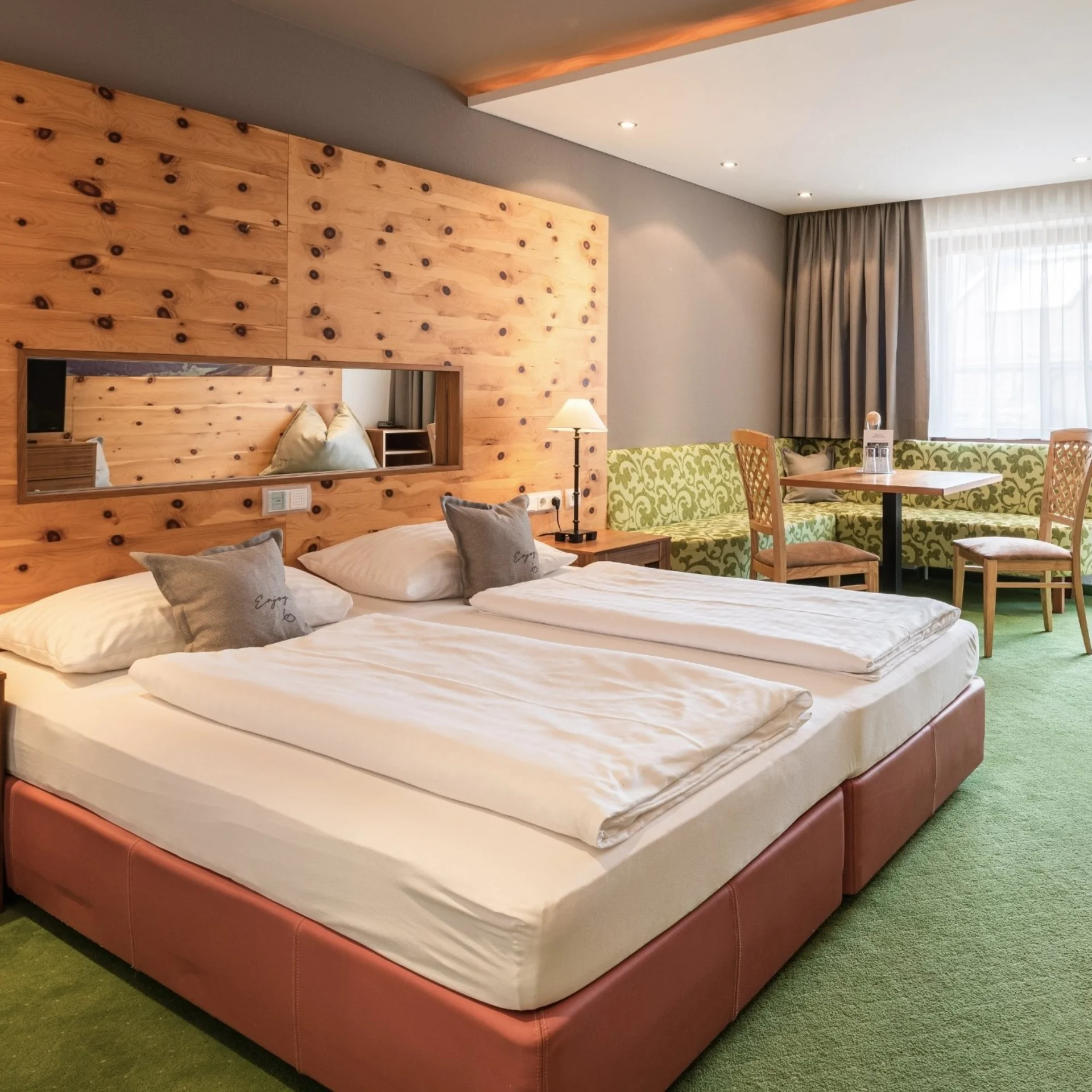 Familienzeit Helles Hotelzimmer mit Doppelbett, Holzwand, Sitzecke und grünem Teppich