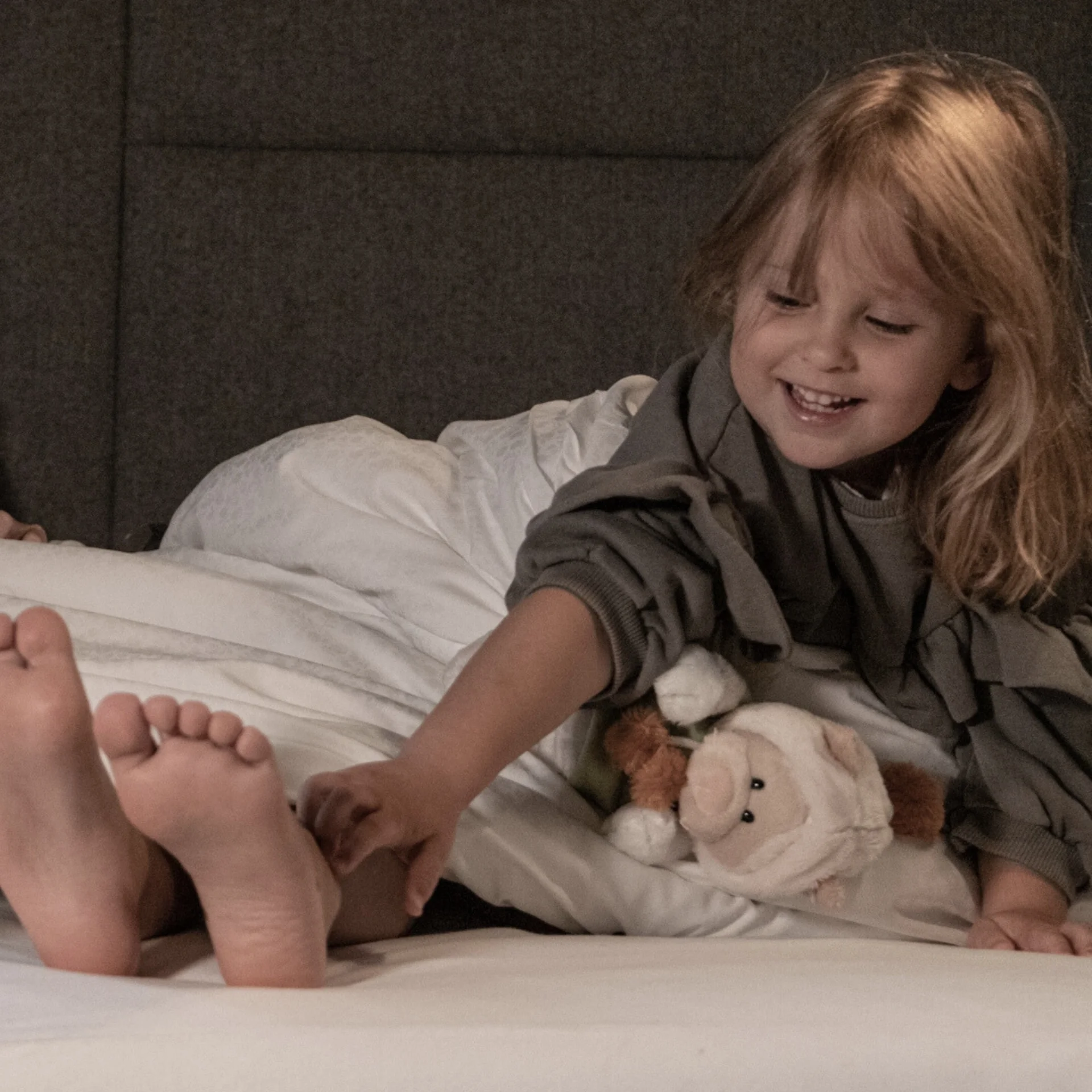Familienzeit Drei Kinder spielen lachend im Bett, zwei unter der Decke, eines mit Kuscheltier
