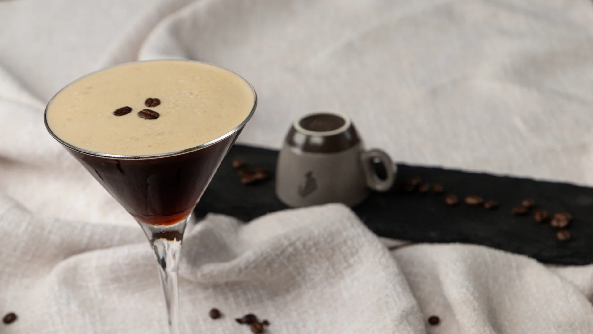 Wohnen Espresso Martini mit Schaum und Kaffeebohnen oben auf, daneben Kaffeebohnen auf Tuch