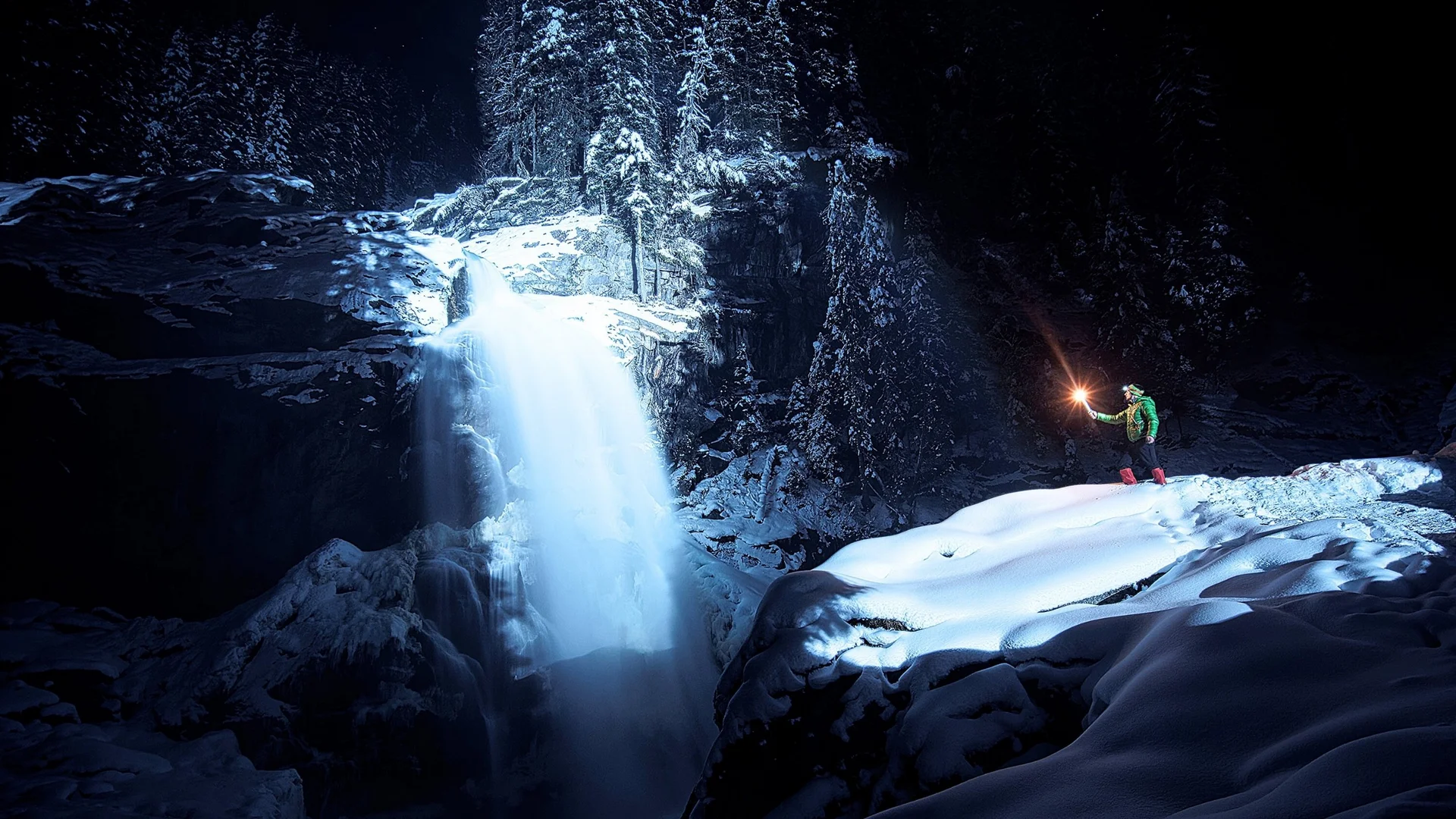 Abseits der Piste Person hält bei Nacht im verschneiten Wald eine Lichtquelle nahe einem Wasserfall