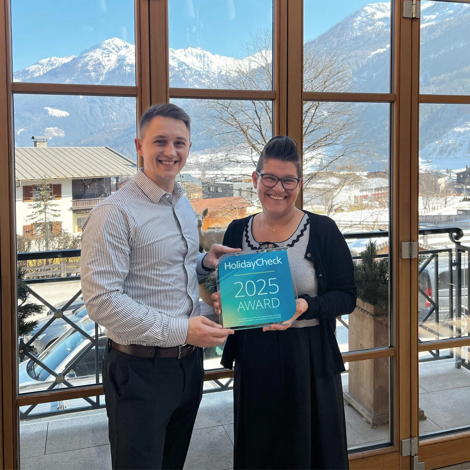 Bewertungen Zwei Personen halten HolidayCheck Award 2025 vor Fenster mit Bergblick