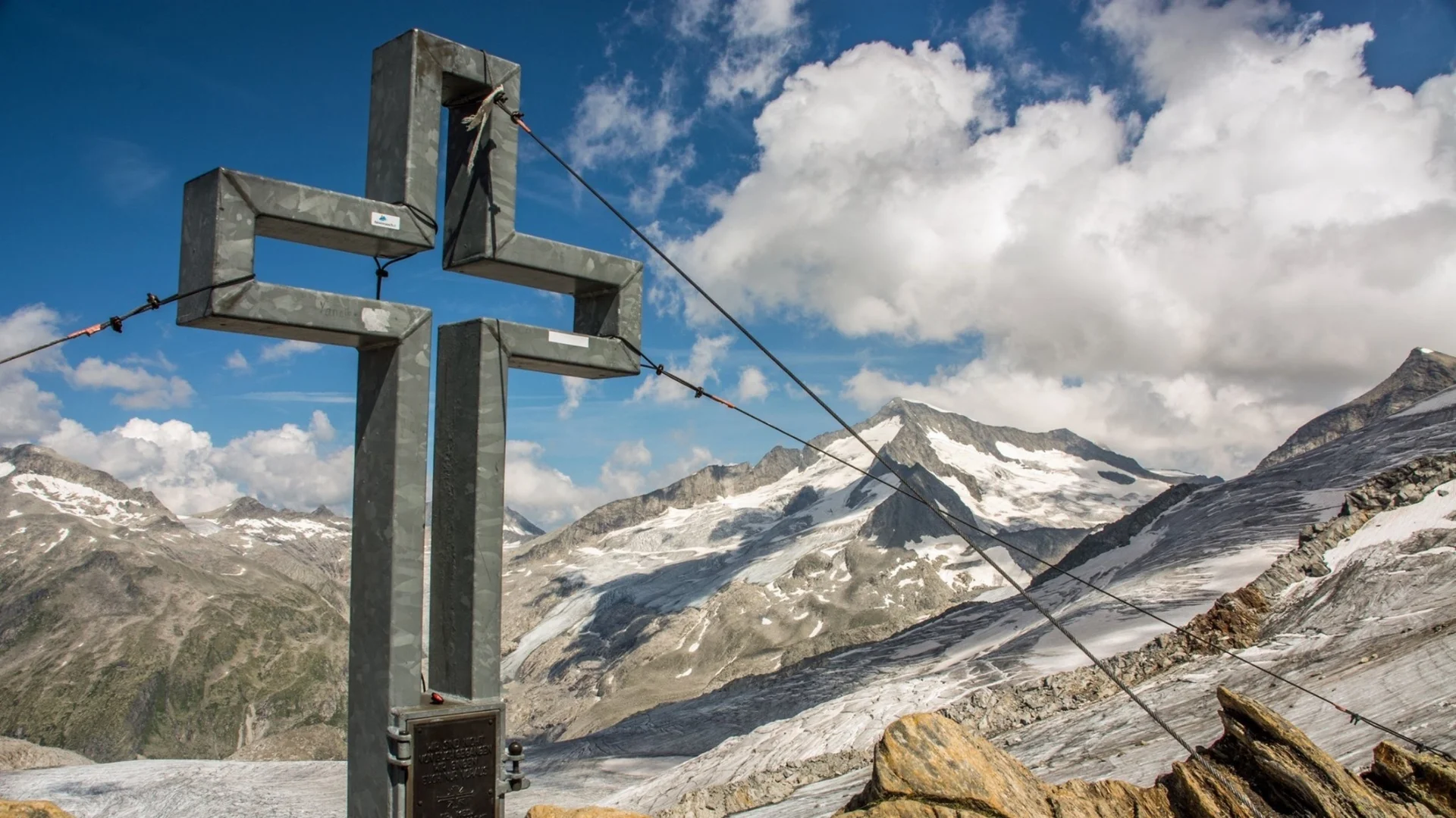 Wanderservice Metallkreuz auf Berggipfel mit schneebedeckten Alpen und Wolken am Himmel