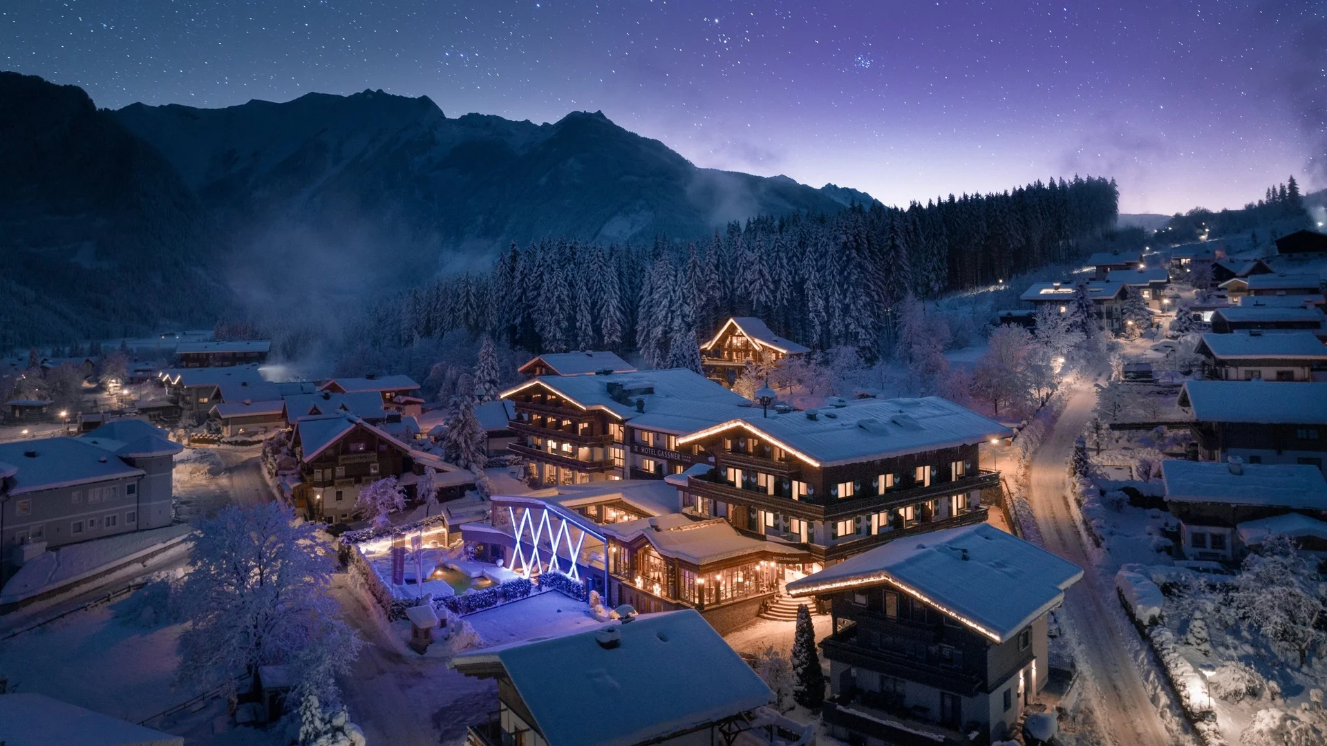 Das Hotel Schneebedecktes Dorf bei Nacht mit beleuchteten Häusern und Bergen im Hintergrund