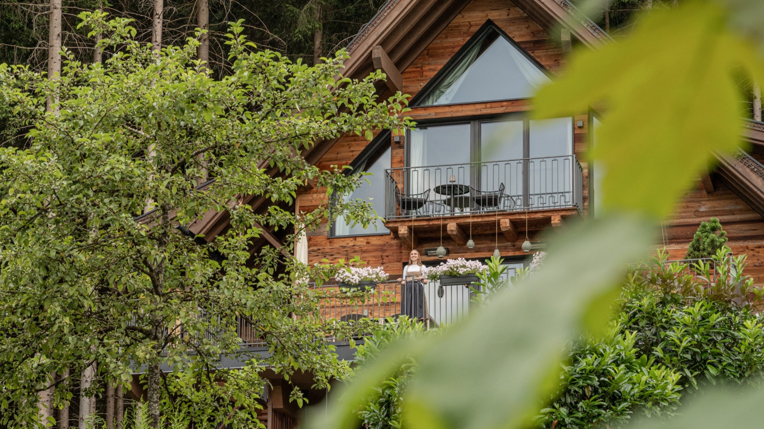 Home Holzchalet mit Balkon und Frau, umgeben von Bäumen und Pflanzen