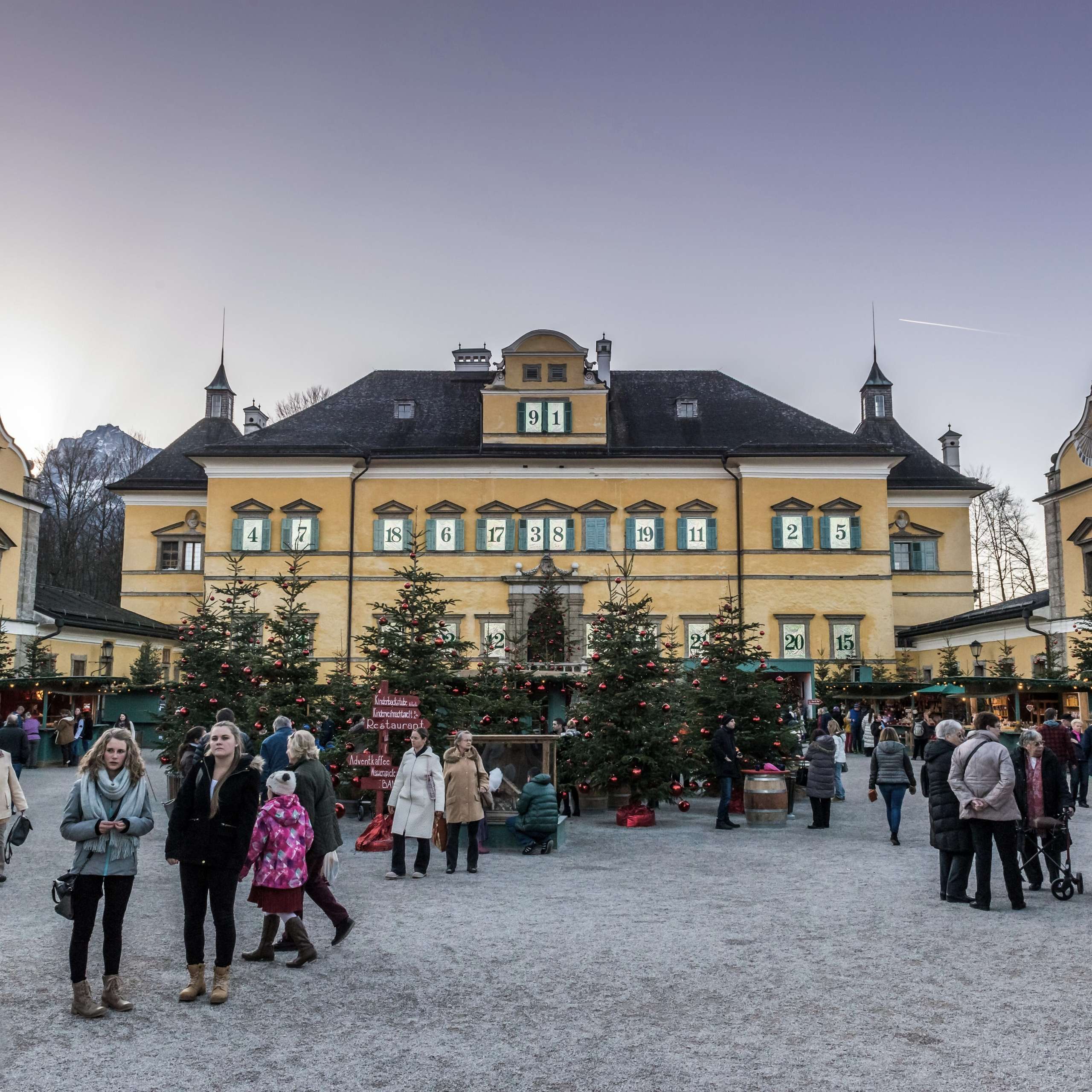 Ausflugstipps Weihnachtsmarkt beim Schloss Hellbrunn in Salzburg mit geschmückten Tannen, Marktständen und Besuchern im Abendlicht