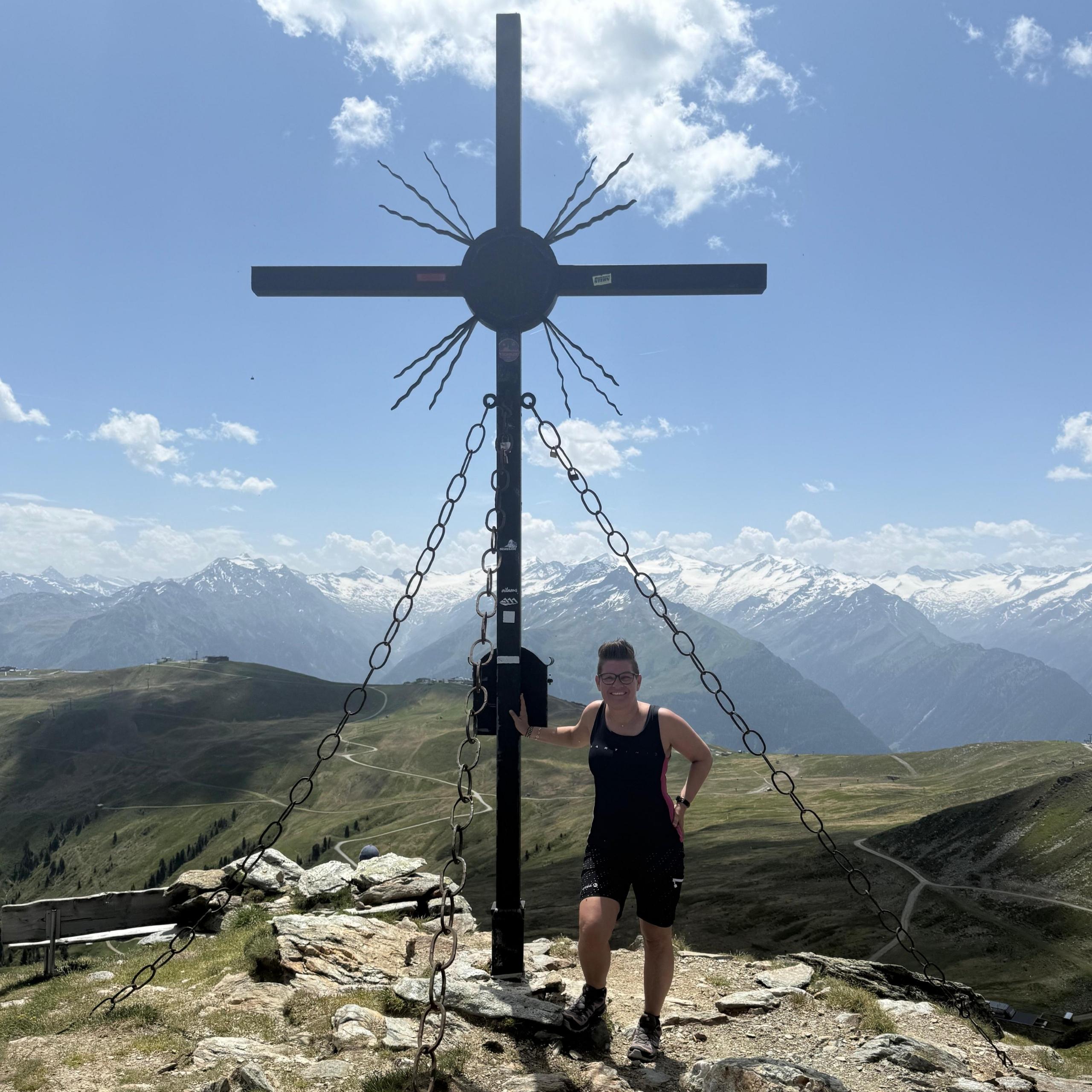 Ausflugstipps Frau steht am Gipfelkreuz vom Frühmesser mit Alpen im Hintergrund an einem sonnigen Tag