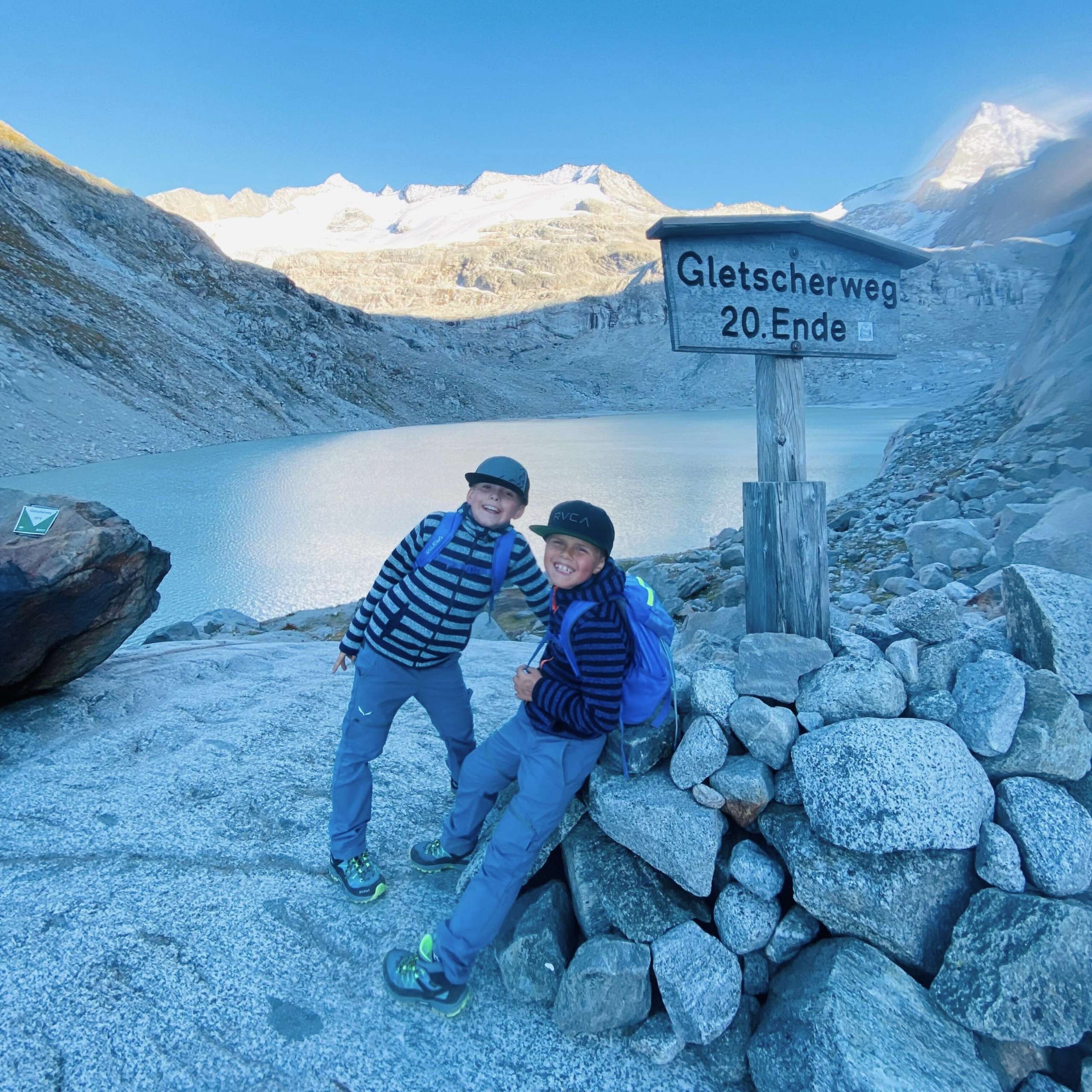 Ausflugstipps Zwei Kinder mit Rucksäcken stehen am Gletscherlehrweg-Schild vor dem vom Gletscher gespeisten Bergsee und schneebedeckten Bergen im Hintergrund im Obersulzbachtal