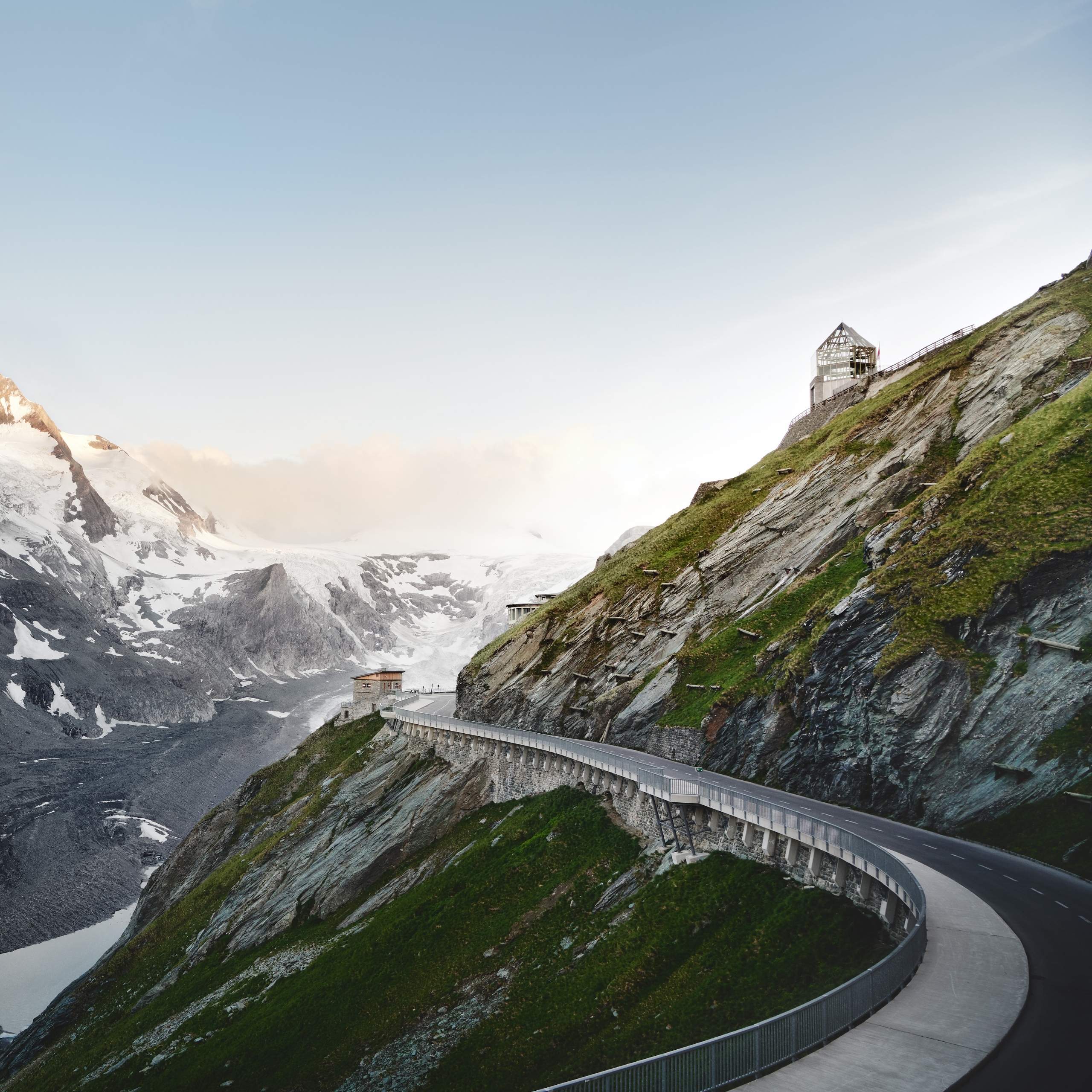 Ausflugstipps Straßenabschnitt der Großglockner Hochalpenstraße entlang einer steiler Felswand mit Schnee bedeckten Gipfeln im Hintergrund