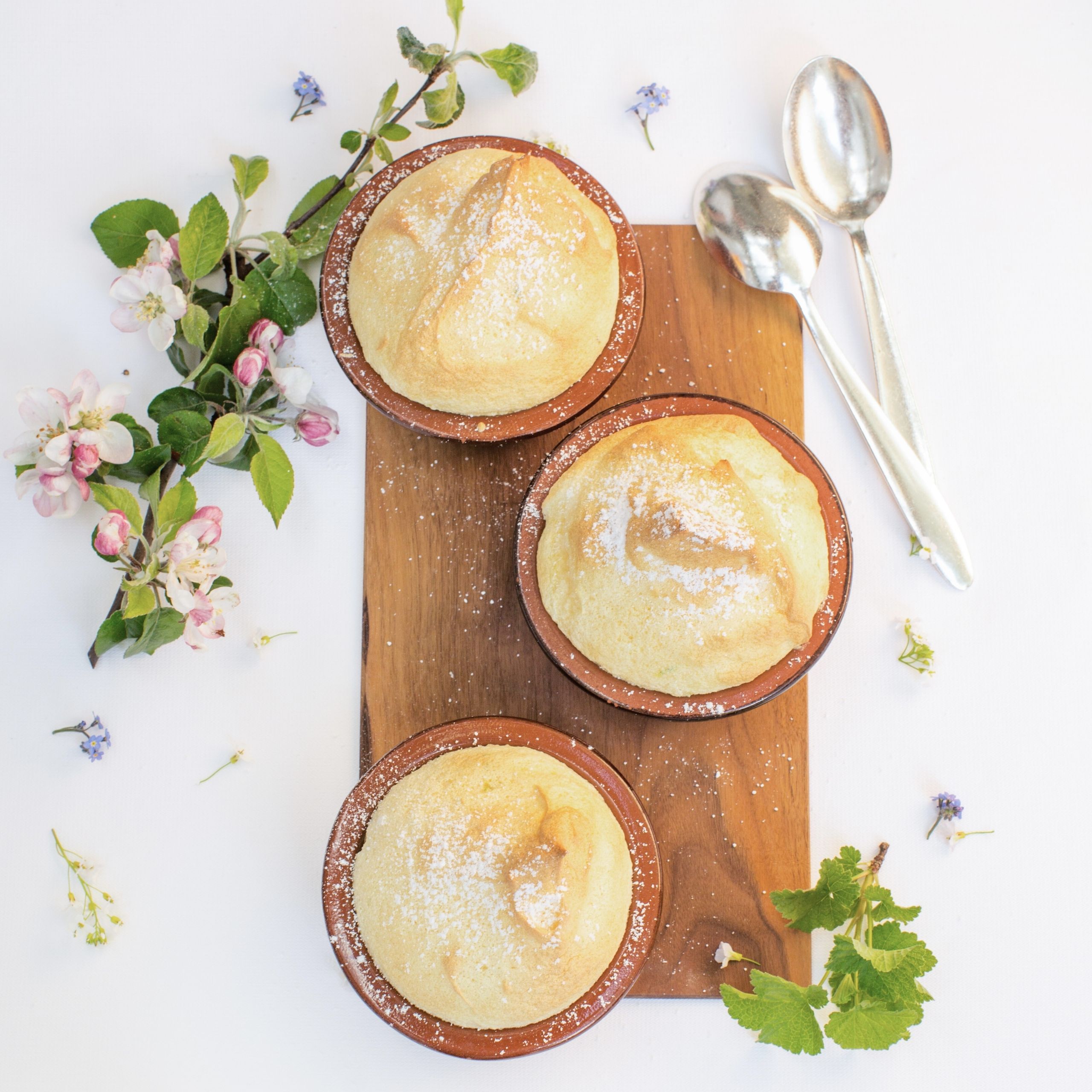 Ausflugstipps Drei Soufflés in braunen Förmchen mit Puderzucker auf Holzbrett, Blumen und Löffel als Dekoration