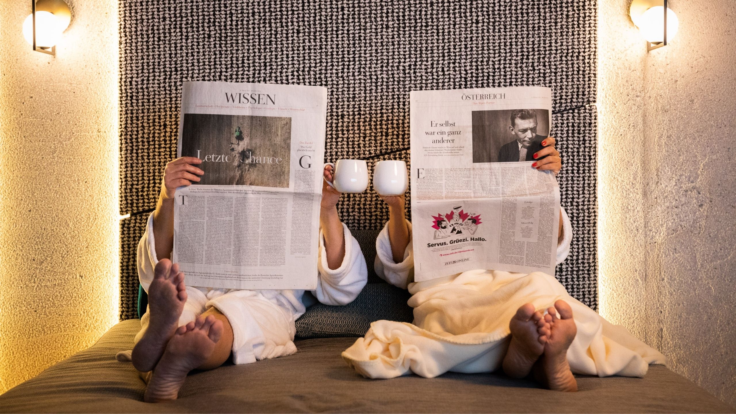 Saunawelt Zwei Personen in Bademänteln lesen Zeitung und stoßen mit Tassen an